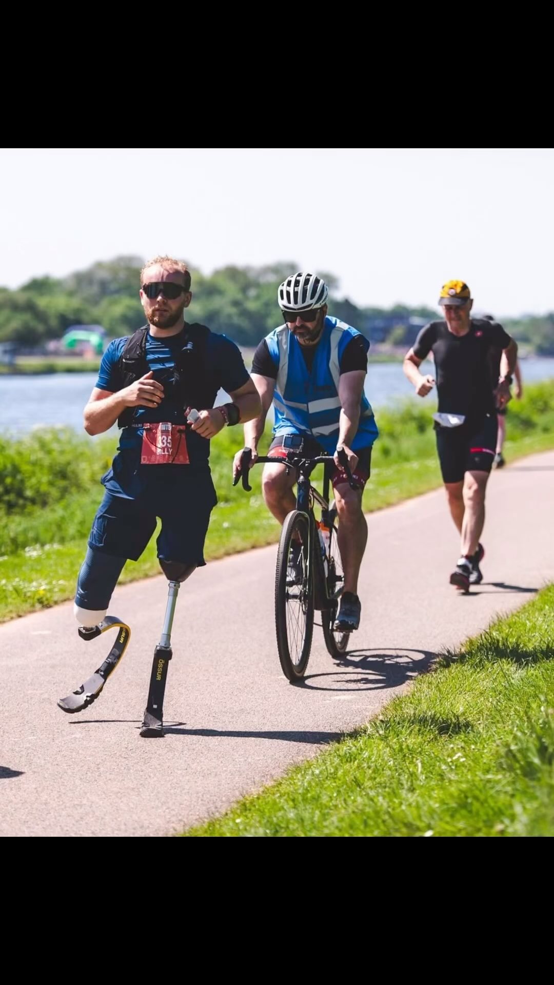 Billy Monger running beside a cyclist and another runner on a lakeside path